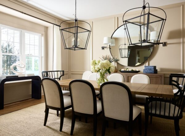Elegant dining room with a wooden table, eight cushioned chairs, two large black lantern pendant lights, a round mirror, sideboard, white flowers in a vase, and natural light from large windows.