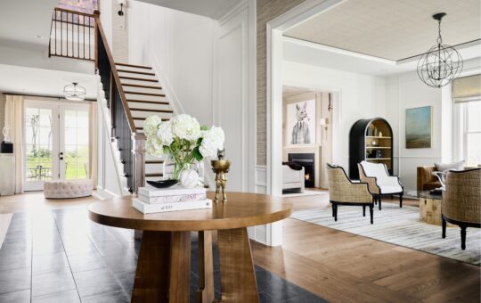 Bright, modern entryway with a round wooden table holding flowers and books, leading to a stylish living room with neutral furniture, art on the walls, and large windows letting in natural light. A staircase is visible to the left.