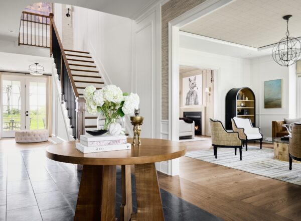 Bright, modern entryway with a round wooden table holding flowers and books, leading to a stylish living room with neutral furniture, art on the walls, and large windows letting in natural light. A staircase is visible to the left.