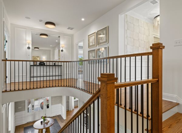 A bright, modern hallway with wooden stairs and black metal railings, leading to an open landing. Light fixtures, framed art, and a view of the lower entryway with a round table and decor are visible.
