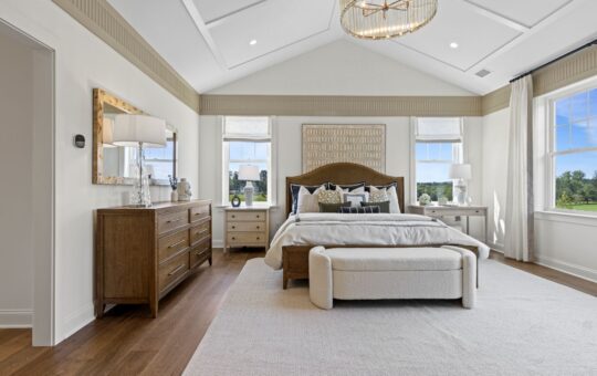Spacious, modern bedroom with a large bed, cushioned bench, wooden dresser, and nightstands. Neutral tones, large windows with views of greenery, white walls, wooden floor, and elegant light fixture.