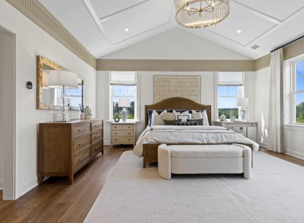 Spacious, modern bedroom with a large bed, cushioned bench, wooden dresser, and nightstands. Neutral tones, large windows with views of greenery, white walls, wooden floor, and elegant light fixture.