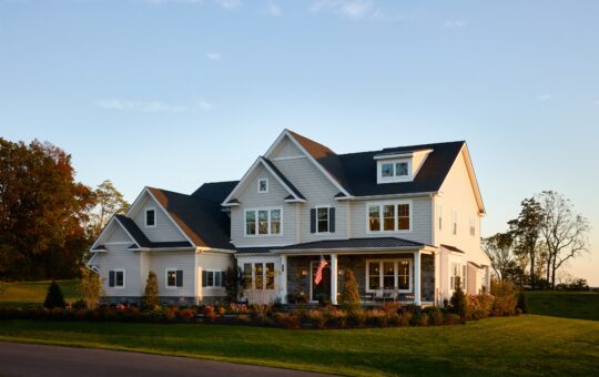 A large two-story suburban house with white siding, multiple gables, and a front porch. An American flag hangs near the entrance, and the home is surrounded by a well-kept lawn and landscaped bushes at sunset.