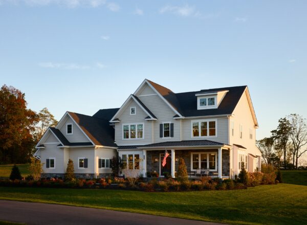 A large two-story suburban house with white siding, multiple gables, and a front porch. An American flag hangs near the entrance, and the home is surrounded by a well-kept lawn and landscaped bushes at sunset.