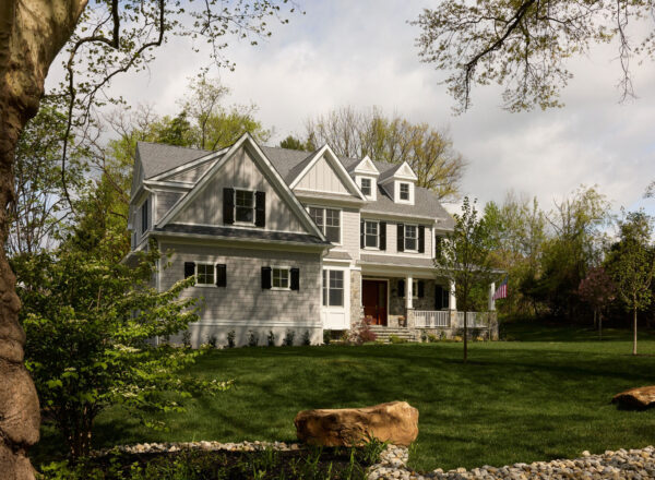 A large white two-story house with multiple gables and black shutters sits surrounded by green grass, trees, and bushes. An American flag hangs on the front porch, and rocks are landscaped in the yard.