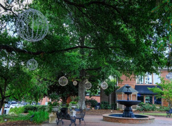 A small brick fountain surrounded by benches and greenery sits under large trees decorated with hanging spherical lights, near a red brick building.