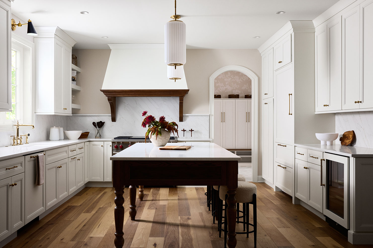 A spacious, elegant kitchen with white cabinets, marble countertops, wood flooring, a large island with stools, gold fixtures, and a central pendant light. A vase with flowers decorates the island.