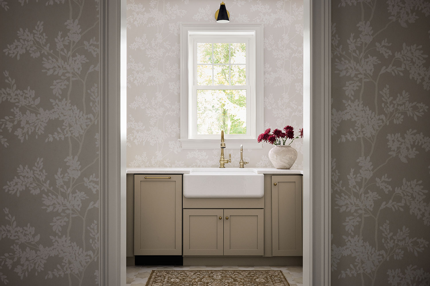 A bright, stylish bathroom with beige cabinets, a white farmhouse sink, gold faucet, and a vase of red flowers on the counter. A window above the sink lets in natural light, and floral wallpaper covers the walls.