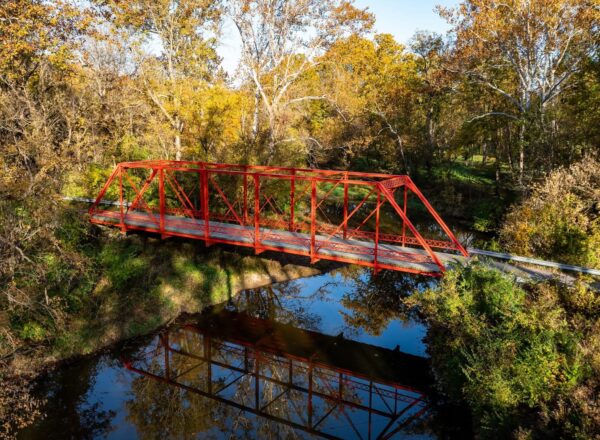 A bright red truss bridge spans over a calm river, surrounded by dense trees with autumn foliage. The bridges reflection is clearly visible in the water below.