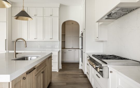 Modern kitchen with white cabinets, stainless steel appliances, a large island with a sink, brass fixtures, pendant lights, light wood flooring, and an arched doorway leading to a pantry.