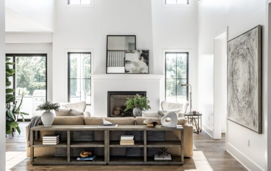 A bright, modern living room with large windows, neutral decor, a beige sofa with built-in shelves, potted plants, a fireplace, abstract art, and wooden floors. Sunlight fills the airy space.