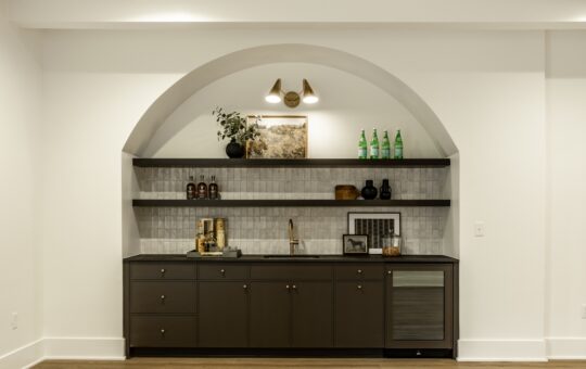 A modern home bar with dark cabinets, open shelves, a small sink, a mini fridge, bottles, decor, and an arched alcove with overhead lighting against a white wall and wood flooring.