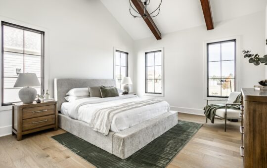 Bright, modern bedroom with a gray upholstered bed, white bedding, wooden nightstand and dresser, green rug, large windows, and exposed wood ceiling beams. Room is decorated in neutral tones with minimalist decor.