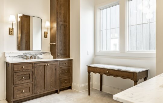 Bright bathroom with wooden cabinets, a marble countertop, a rectangular mirror with wall sconces, a small wooden vanity table by two large windows, and beige tiled floor.