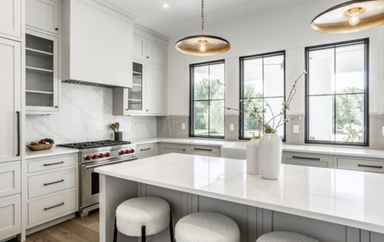 Modern kitchen with white cabinets, marble countertops, a gas stove with red knobs, two pendant lights, large windows, and a white island with two decorative vases and upholstered stools.