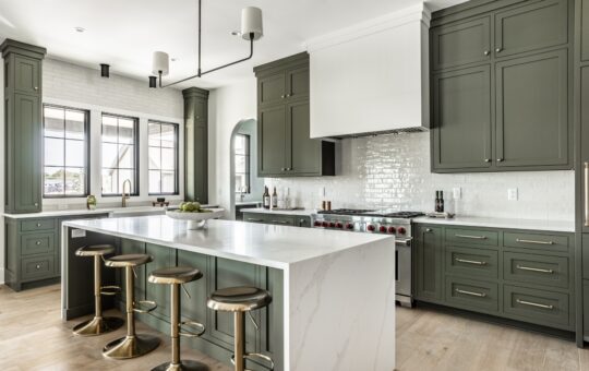 Modern kitchen with green cabinets, white marble countertops, a large island with gold stools, stainless steel appliances, and large windows letting in natural light.