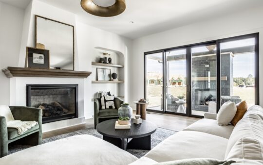 Modern living room with a corner fireplace, built-in shelves, green armchairs, a white sectional sofa, round black coffee table, large windows, and a view of a patio and outdoors. Decor is neutral and contemporary.