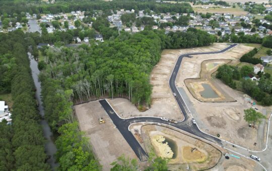 Aerial view of a partially developed residential area with newly paved roads, construction vehicles, patches of trees, small ponds, and surrounding neighborhoods with houses and greenery.