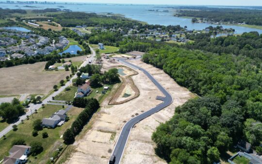 Aerial view of a newly paved road winding through a cleared area surrounded by homes, trees, and water, with a distant city skyline and coastline in the background.