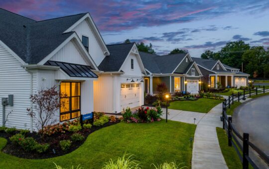 Row of modern, single-story suburban homes with manicured lawns and landscaped gardens at sunset. Warm lights glow from windows, and a curved sidewalk runs along the houses.