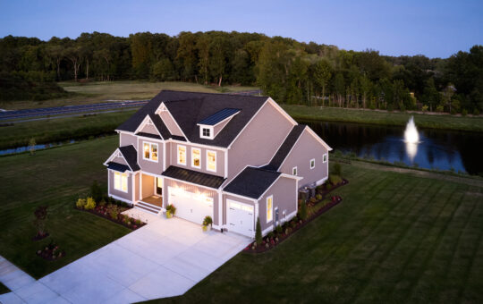 A large two-story house with a well-kept lawn, driveway, and lit windows at dusk. The house sits near a small pond with a fountain, surrounded by trees and greenery in the background.