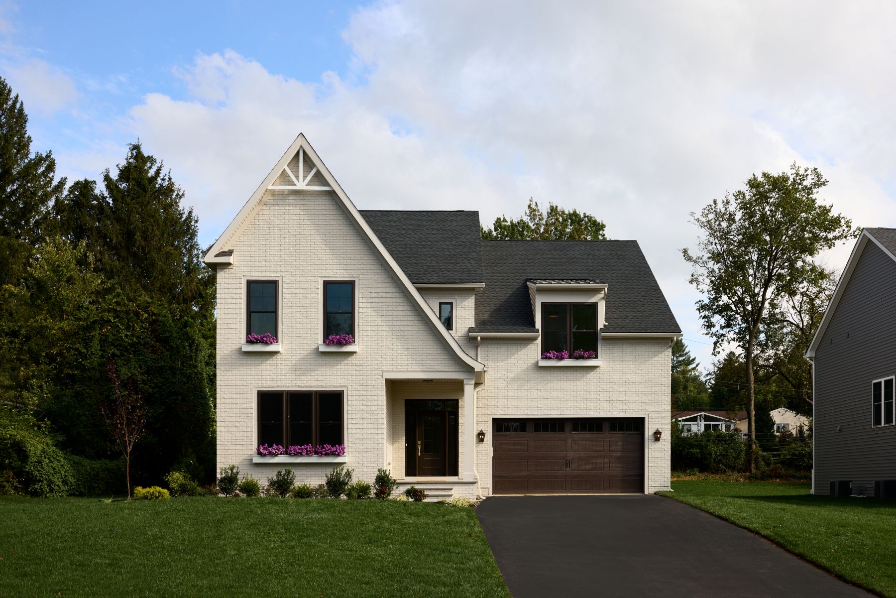 A modern two-story white brick house with a dark gray roof, brown garage door, and flower boxes under the windows. The house sits on a green lawn with a paved driveway and trees in the background.