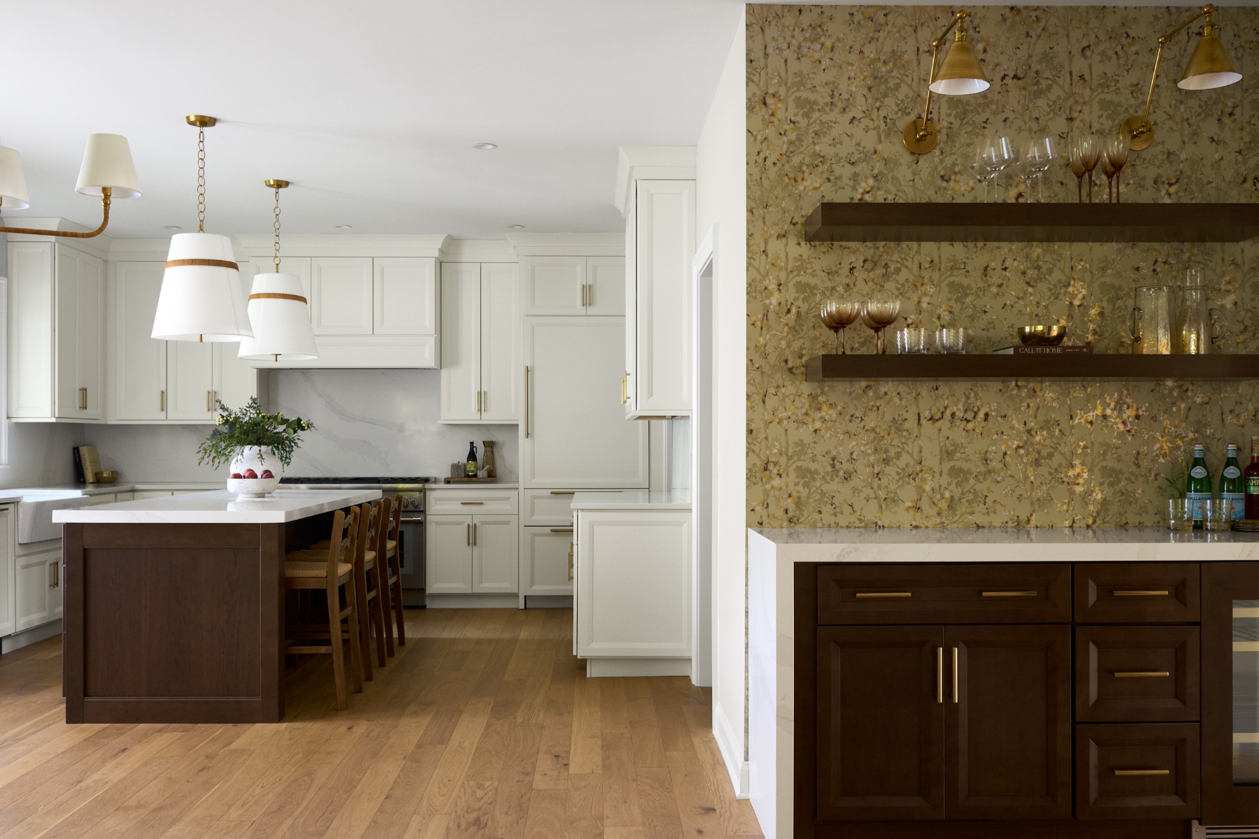 A modern kitchen with white cabinets, a dark wood island with bar stools, gold light fixtures, and a side bar area featuring open shelves, glassware, and a decorative gold-patterned backsplash.