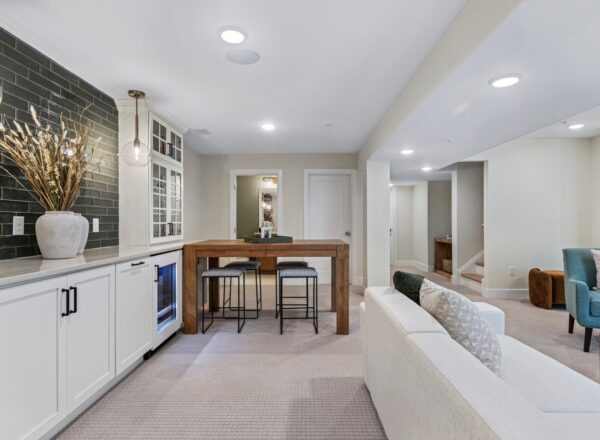 Modern open-concept living area with a small bar featuring a wooden table and two stools, white cabinetry, a backsplash with dark tiles, a white sofa, and a blue armchair in a bright, neutral-toned space.
