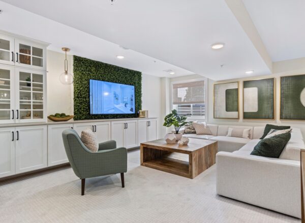 A modern living room with a white sectional sofa, green armchair, wooden coffee table, and a large TV mounted on a wall with green foliage. White cabinets, abstract artwork, and plants add to the bright, cozy decor.