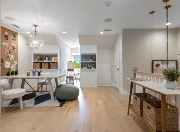A bright, modern creative workspace with a large white table, round stools, a pinboard with photos, shelves with baskets and art supplies, wooden floors, and pendant lights, all in a neutral-toned room.
