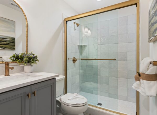 A modern bathroom with a glass shower enclosure, gold fixtures, gray vanity with a marble countertop, a potted plant, wall art, and neatly rolled towels on a rack.