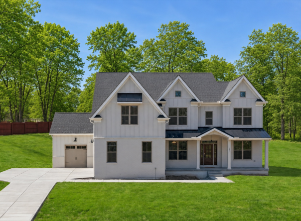 A modern two-story white house with black trim, large front porch, attached garage, and a well-kept lawn, surrounded by green trees under a clear blue sky.
