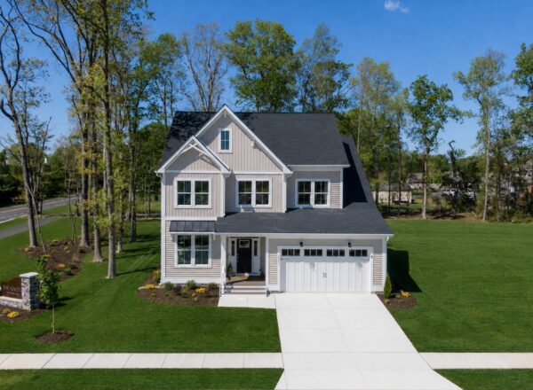 A two-story beige house with white trim, attached garage, large driveway, neat lawn, and surrounded by trees under a clear blue sky.