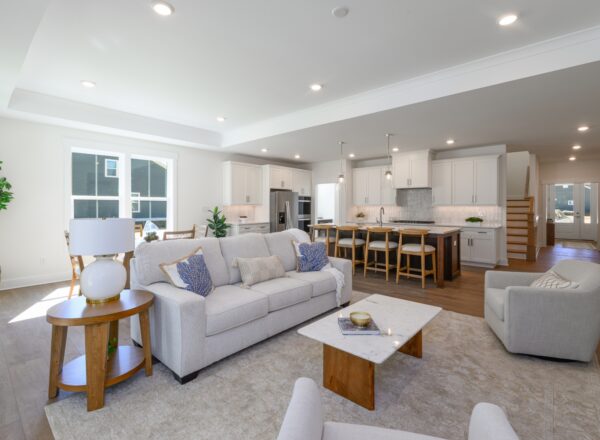 Modern open-concept living room and kitchen with white cabinetry, light countertops, a large island with bar stools, and neutral-toned furnishings. Natural light streams in through large windows, brightening the space.