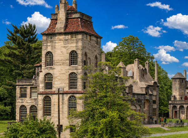 A unique, castle-like stone building with large windows and a red-tiled roof stands among green trees under a blue sky with scattered clouds.