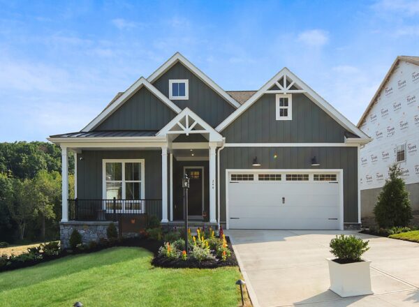 A one-story dark gray house with a two-car garage and a small covered front porch, next to a house under construction.